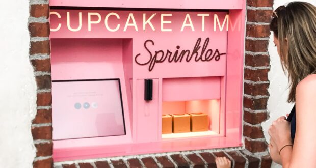 A woman stands in front of a pink Sprinkles cupcake ATM built into a brick wall. Three cupcake boxes are visible inside the machine’s open window.