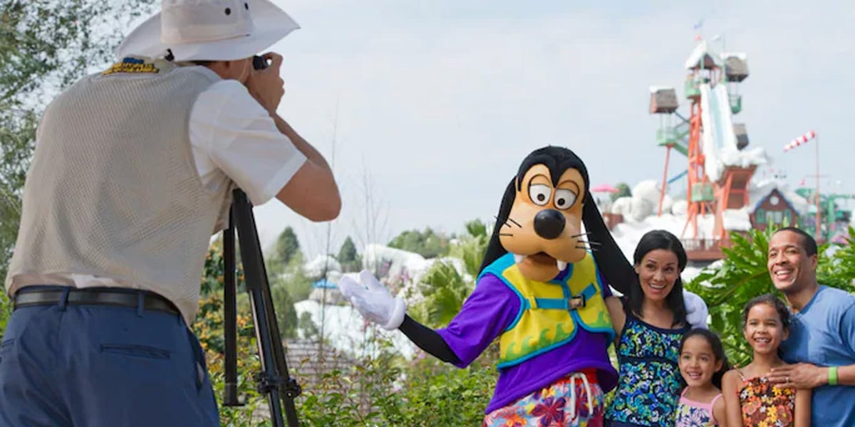 A photographer captures a smiling family posing with Goofy at an amusement park, with water slides and trees in the background—it's a perfect Memory Maker moment.