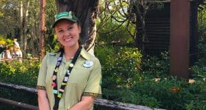 A cheerful Cast Member in classic green attire greets guests under lush trees at a Disney park, ready to assist with magic.