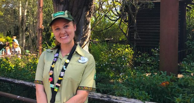 A cheerful Cast Member in classic green attire greets guests under lush trees at a Disney park, ready to assist with magic.