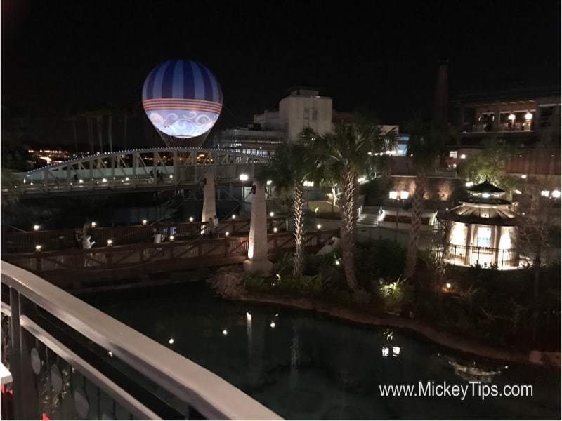 Disney Springs at night with a glowing bridge, soaring striped balloon, palm trees, and Planet Hollywood shining in the background.