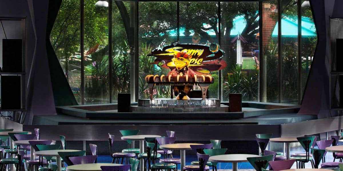 A colorful indoor stage with a neon sign reading "Sonny Eclipse" is set for a performance. The stage features a futuristic design, with tables and chairs arranged in the foreground. Large windows showcase a view of green trees and outdoor structures.
