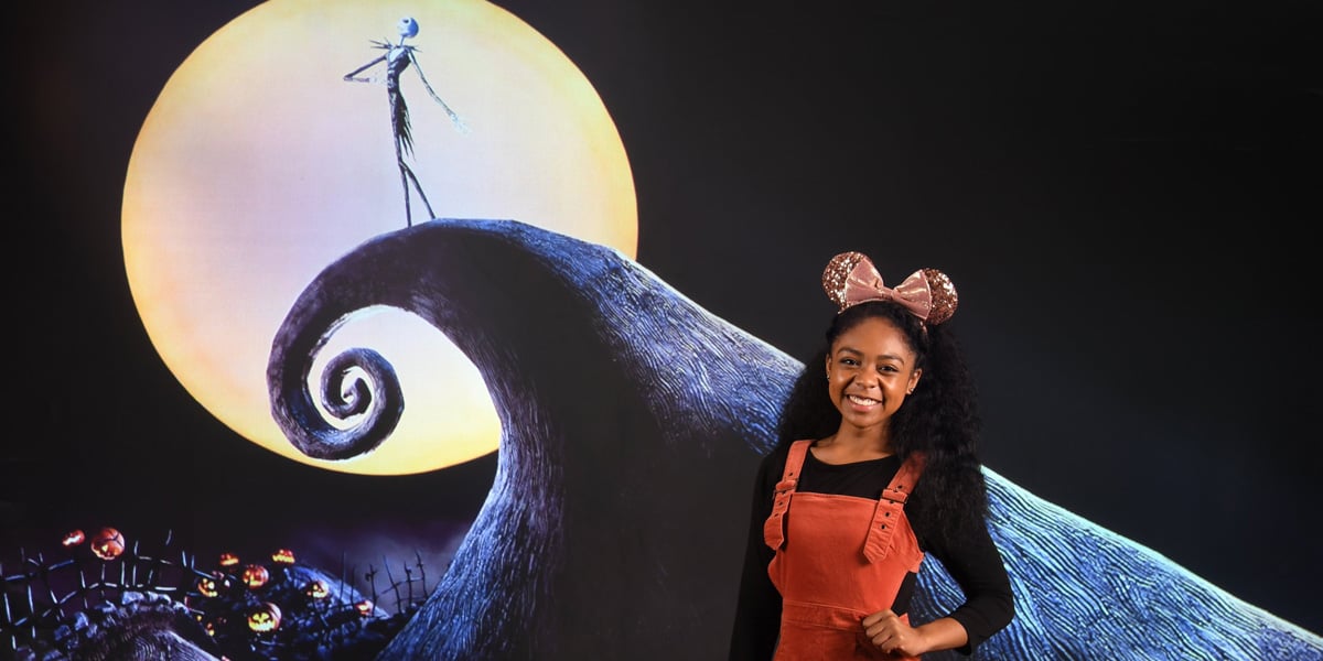 A smiling woman wearing Minnie Mouse ears and an orange overall dress stands in front of a Memory Maker backdrop featuring Jack Skellington from "The Nightmare Before Christmas" silhouetted against a large moon.