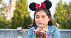 A young girl wearing Minnie Mouse ears and a red bow blows colorful confetti from her hands while smiling outdoors, capturing a Memory Maker moment. Green trees and a blurred castle are visible in the background.