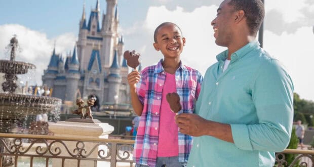 A smiling child and adult enjoy Mickey Mouse-shaped ice cream bars included with the Disney Dining Plan in front of a castle at a theme park, with a fountain and blue sky in the background.
