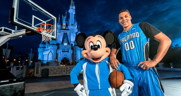 Mickey Mouse and an Orlando Magic player smile by a basketball hoop, Disney castle glowing behind them at dusk in Walt Disney World.