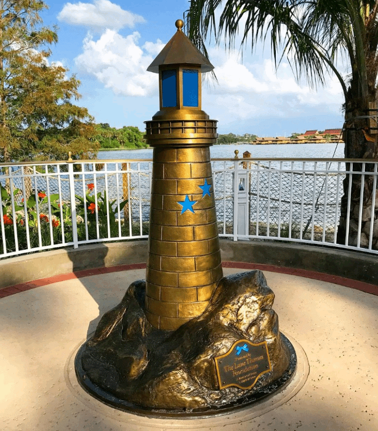 Bronze lighthouse with blue stars by a lakeside, palm trees, and white fence, honoring Lane Graves at Disney World.