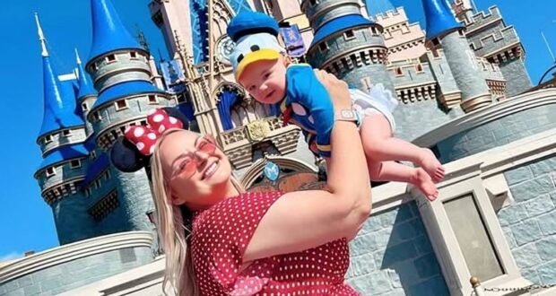 A smiling woman in Minnie Mouse ears and a red polka-dot dress holds up her baby, dressed as Donald Duck, in front of a blue and pink Disney castle on a sunny day—capturing one of many Mother-Son Disney World Activities.