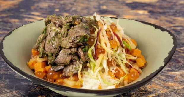 A bowl of sliced beef topped with green herbs, served with shredded cabbage, diced sweet potatoes, and small round brown pearls on the side. The dish is presented in a light green bowl against a textured background.