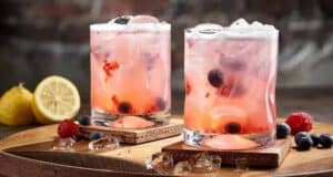 Two glasses of pink, berry-filled cocktails with ice, garnished with blueberries and raspberries, sit on wooden coasters—evoking the vibrant drinks served at Disney Springs Restaurants. Nearby, a halved lemon and scattered berries complete the inviting scene.