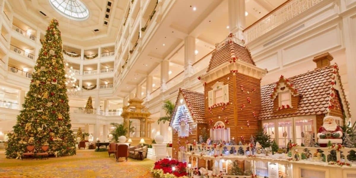 A grand hotel lobby beautifully decorated for Christmas. A large, ornate Christmas tree stands beside an elaborate gingerbread house adorned with festive decorations, including Santa Claus figures and holiday lights. The space is warm and inviting.