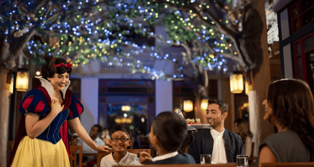 A person dressed as a fairy-tale character interacts with a family dining in a restaurant. The setting is decorated with twinkling lights and trees. The family includes two adults and two children, and there's a server holding a tray.