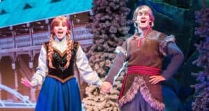 Two performers dressed as characters from a winter-themed fairy tale, one in a blue dress with braids, and the other in a tunic with fur trim. They hold hands, singing on a snowy stage with a backdrop of a snowy village.