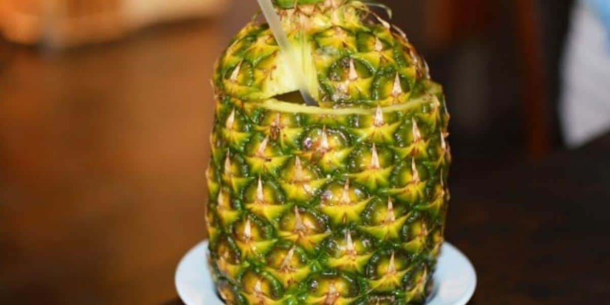 A fresh pineapple with the top cut off and hollowed out, filled with a beverage. A straw is inserted for drinking, and the pineapple is placed on a white plate against a blurred background.