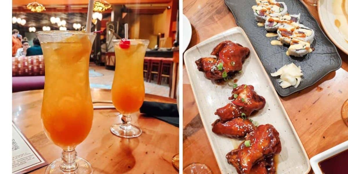 Two orange cocktails with cherries on a wooden table, alongside a plate of glazed chicken wings garnished with green onions, and a slate with sushi rolls drizzled with mayonnaise, ginger, and wasabi, inside a cozy restaurant.