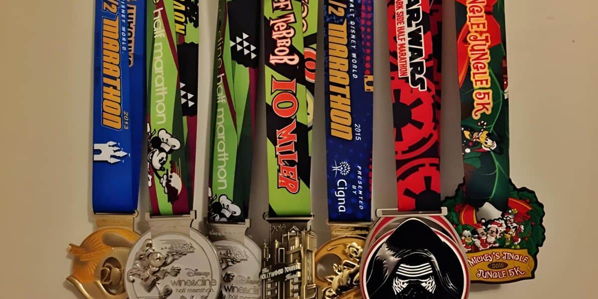 A collection of colorful race medals hanging in a row. Each medal features unique designs and event names, including marathons, a 10-miler, and holiday-themed races. Disney characters are pictured on some of the ribbons.