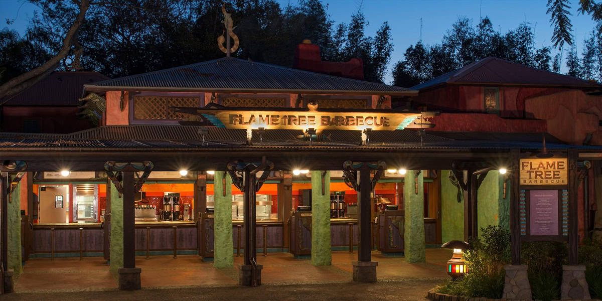 A warmly lit restaurant entrance at dusk displays a sign reading "Flame Tree Barbecue." Wooden columns and colorful decor frame its open service windows, inviting guests to sample signature Flame Tree Barbecue flavors beneath silhouetted trees.