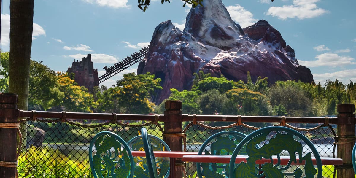 A roller coaster climbs a snowy mountain in the distance, surrounded by lush trees. In the foreground, colorful decorative chairs and a table from Flame Tree Barbecue sit by a fence overlooking the scenic view under a blue sky.