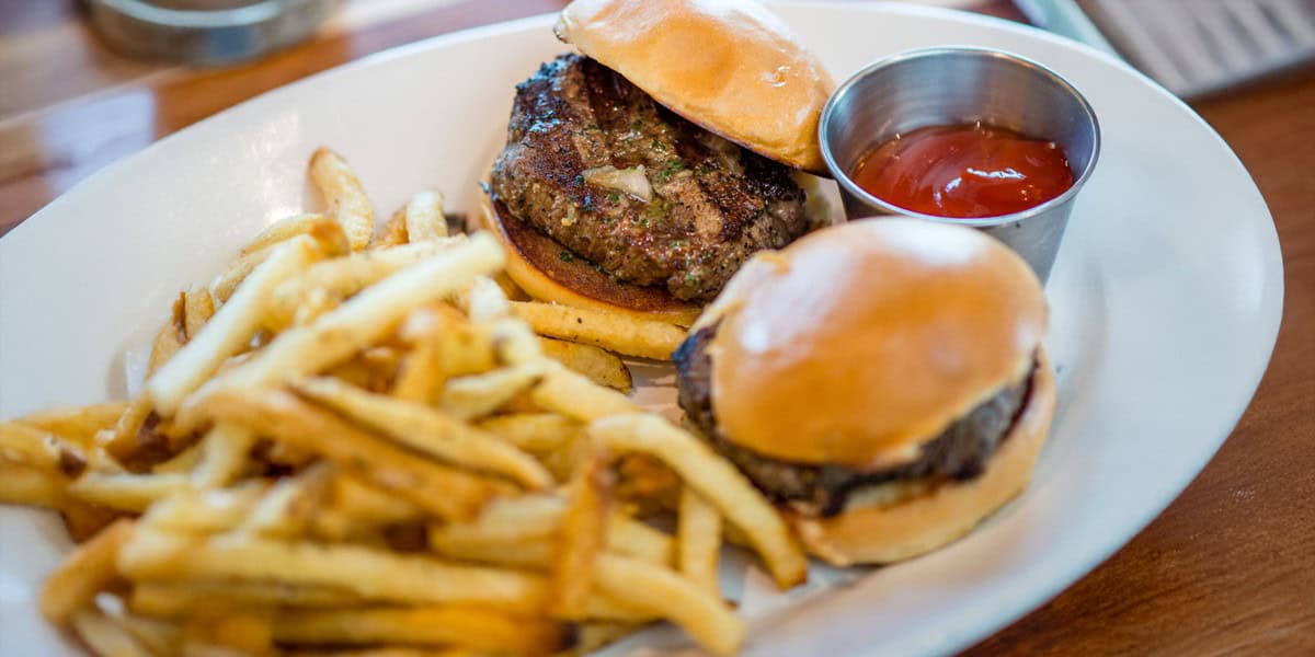 A white plate with two small burgers, a serving of French fries, and a metal cup of ketchup is set on a wooden table at The Boathouse Disney Springs.