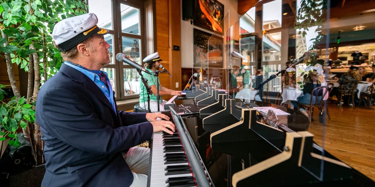 At The Boathouse Disney Springs, a man in a captain's hat plays keyboard and sings into a microphone in a lively restaurant, separated from diners by a clear divider. People in the background also wear captain's hats and sit at tables.