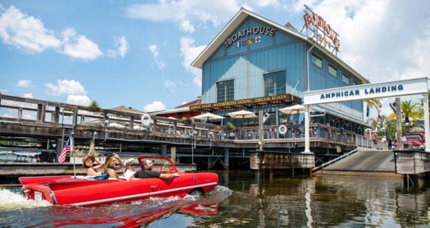 A red amphibious car with three people glides on water near The Boathouse Disney Springs; the blue restaurant features a large sign and a dock labeled "Amphicar Landing" beneath a sunny sky.