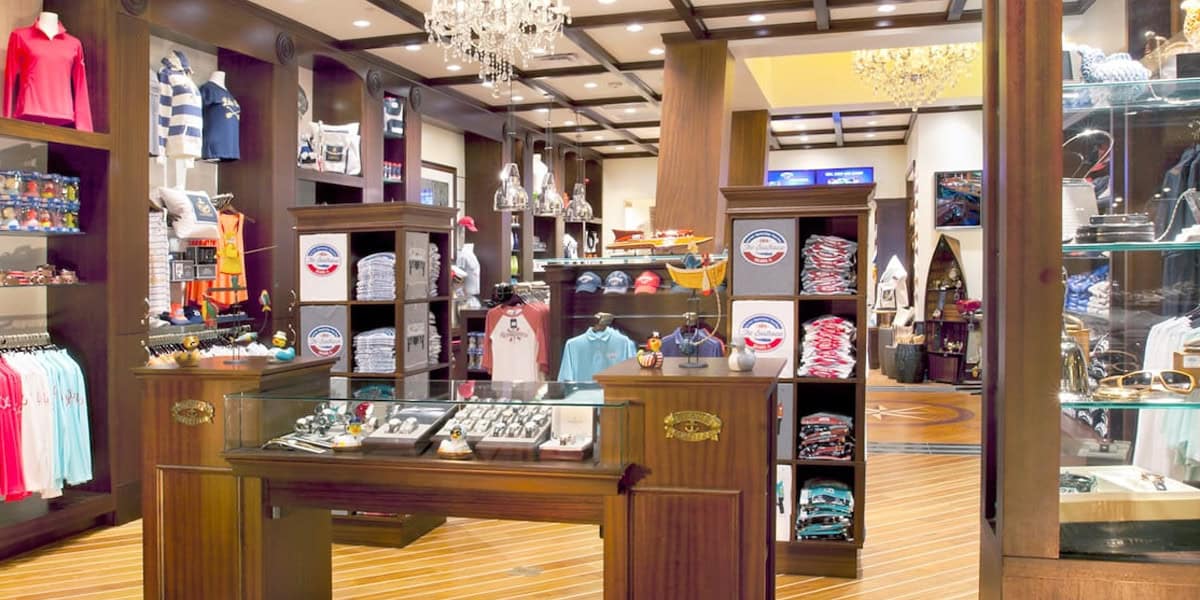 A brightly-lit clothing and accessories store at The Boathouse Disney Springs, with wooden shelves and display cases, featuring neatly arranged shirts, hats, watches, sunglasses, and other merchandise under elegant chandeliers.