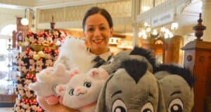 A person smiling while holding several plush toys, including characters resembling a white cat and a gray donkey. The setting appears to be a store with more plush toys displayed in the background.