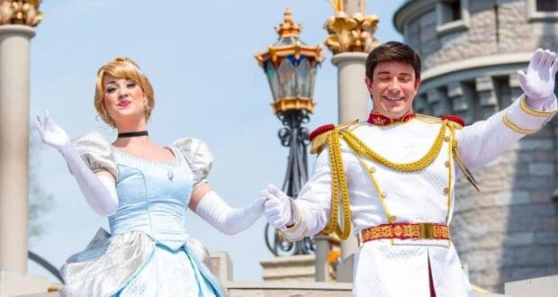 A woman in a blue gown and a man in a white, gold-trimmed outfit wave cheerfully in front of a castle. They are both smiling and posing in a welcoming manner. The setting is bright and sunny.
