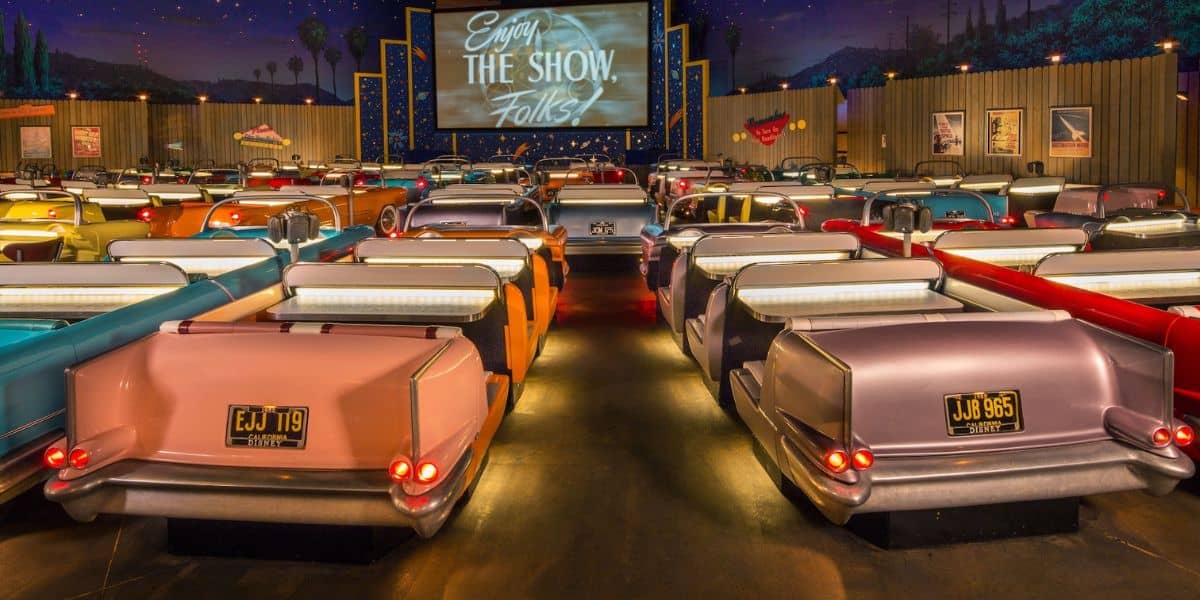 Rows of vintage car-shaped booths inside a retro-themed drive-in theater. A large screen displays "Enjoy the Show, Folks!" The atmosphere is cozy with dim lighting and starry decorations on the ceiling.