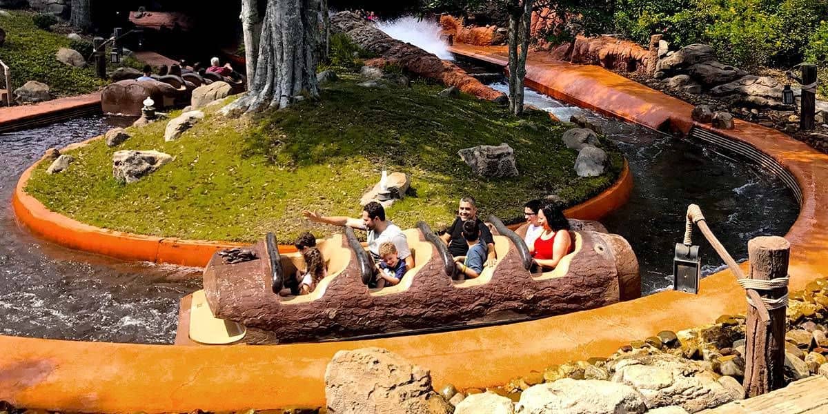 A group of people enjoy a ride in a log-shaped boat on a water theme park attraction. The ride winds through a green, rocky landscape with splashes of water creating an exciting atmosphere.