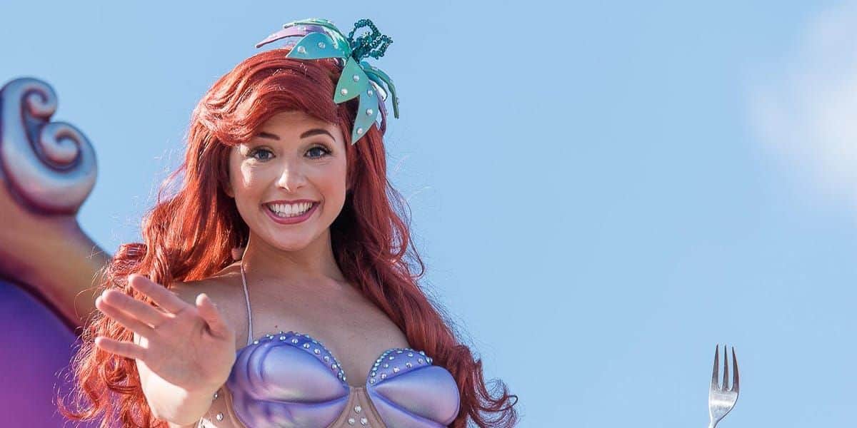 A woman dressed as a mermaid waves happily. She has long red hair, a shell-like top, and a hair accessory with seashells. She holds a fork in one hand against a bright blue sky.