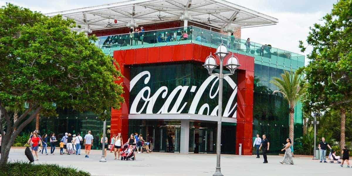 Modern glass building with a large Coca-Cola logo. People are walking around outside. Trees and a street lamp are visible in the foreground.