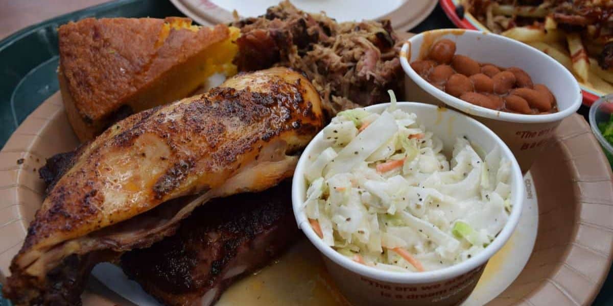 A plate of food featuring roasted chicken, pulled pork, cornbread, baked beans, and a cup of coleslaw.