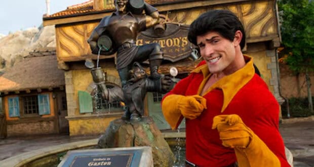 A man dressed as Gaston from "Beauty and the Beast" poses and smiles in front of a bronze Gaston statue holding barrels, outside Gaston’s Tavern—a popular Magic Kingdom dining spot in the theme park.