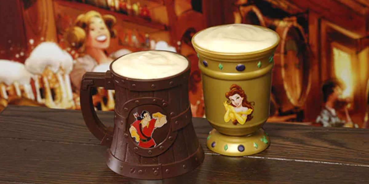 Two novelty mugs filled with frothy drinks sit on a wooden table—perfect for Magic Kingdom dining. One mug features Gaston and is shaped like a barrel, while the other is gold with gem accents and Belle from "Beauty and the Beast.