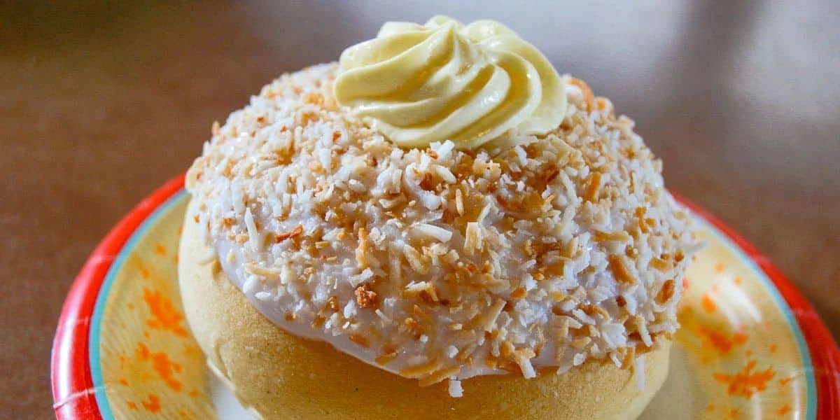 A pastry topped with toasted coconut flakes, white icing, and a swirl of yellow cream sits on a decorative plate with red and yellow accents.