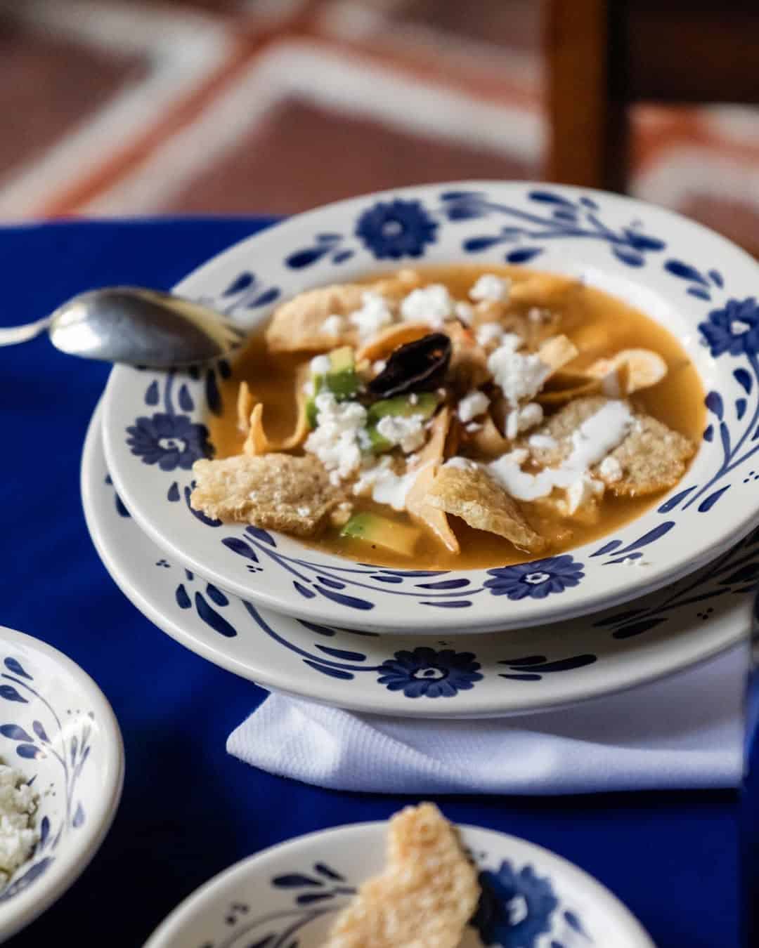 A bowl of soup topped with tortilla chips, cheese, and sour cream sits on a blue floral-patterned plate—reminiscent of the Best Disney World Appetizers—with a spoon beside it and a blue tablecloth underneath.
