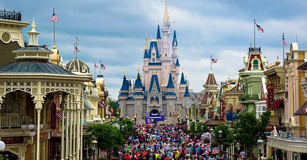 Disney fans flood Main Street toward Cinderella Castle as a Cast Member greets guests under overcast skies in Magic Kingdom.