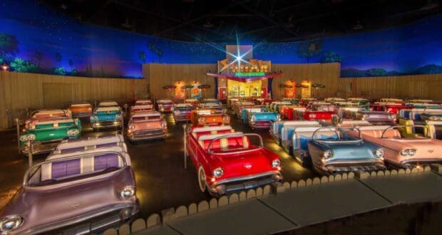 A theater with seating arranged to resemble vintage cars from the 1950s, facing a screen. The room is dimly lit, with a night sky backdrop including stars and palm trees. The decor and colors create a retro, nostalgic ambiance.