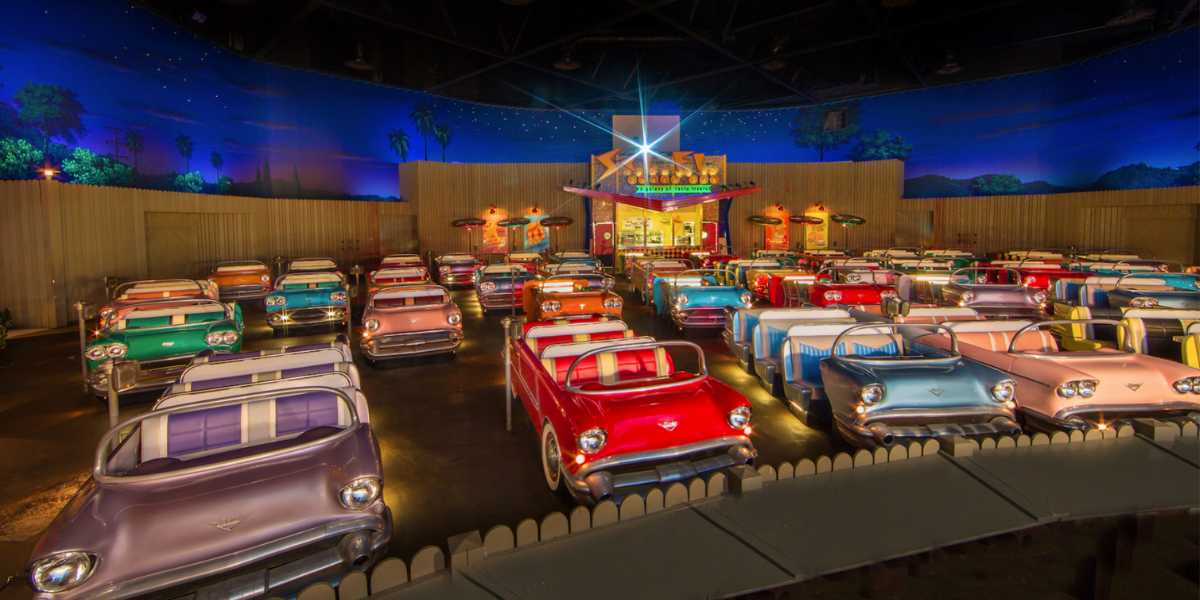 A theater with seating arranged to resemble vintage cars from the 1950s, facing a screen. The room is dimly lit, with a night sky backdrop including stars and palm trees. The decor and colors create a retro, nostalgic ambiance.