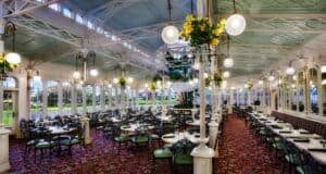 A spacious, elegant dining area with large windows, ornate light fixtures, and green-accented chairs. The ceiling is high with white and blue details, and there are floral arrangements hanging from pillars.