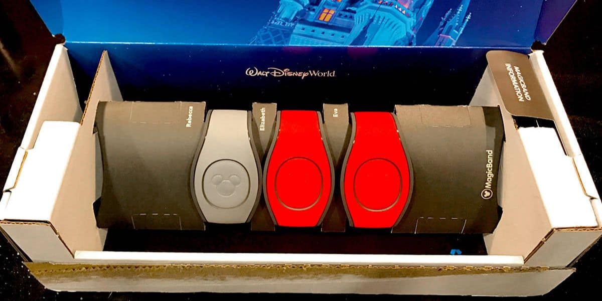 A box containing four Disney MagicBands in different colors: gray, red, and black. The bands are nestled in a branded Disney World package, set against a backdrop featuring a lit-up Disney castle.