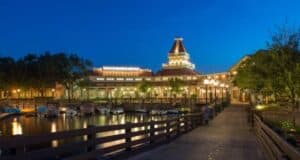 A waterfront view at dusk featuring a well-lit building with a tower and several boats docked along the water. Trees and lampposts line the path leading to the building, creating a serene and inviting atmosphere.