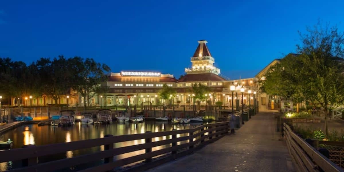 A waterfront view at dusk featuring a well-lit building with a tower and several boats docked along the water. Trees and lampposts line the path leading to the building, creating a serene and inviting atmosphere.