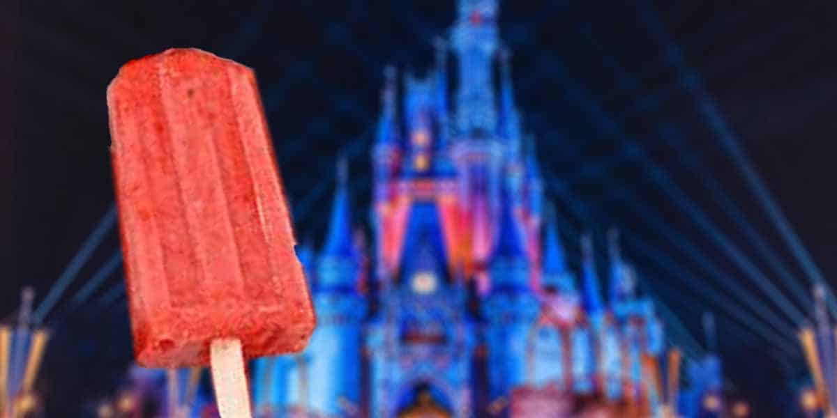 A red popsicle held up in front of a brightly lit, colorful castle at night highlights the magic of Sugar-Free Desserts at Walt Disney World, with blue and purple lights illuminating the enchanting background.