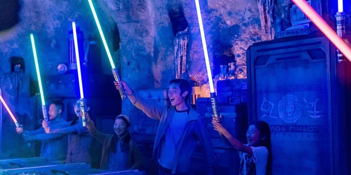 A group of four people joyfully hold up colorful lightsabers in a dimly lit, immersive theme park setting, surrounded by futuristic decor and glowing blue and green lights.