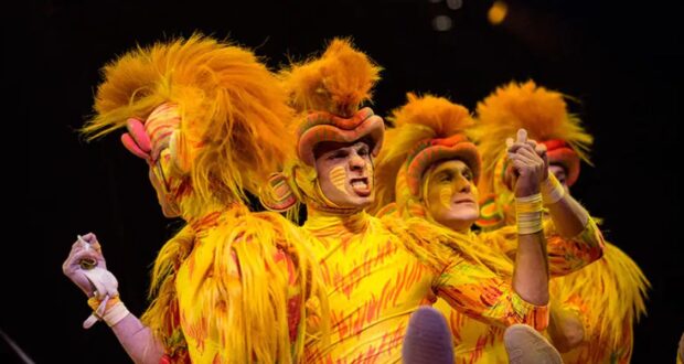 Four performers in vibrant lion costumes with orange and yellow fur and face paint, energetically pose on stage. The background is dark, highlighting their expressive, theatrical gestures.
