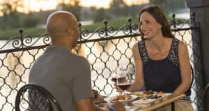 A man and woman sit at an outdoor restaurant table by a decorative railing, sharing Epcot alcoholic drinks, smiling and engaged in conversation as the sun sets over the water in the background.