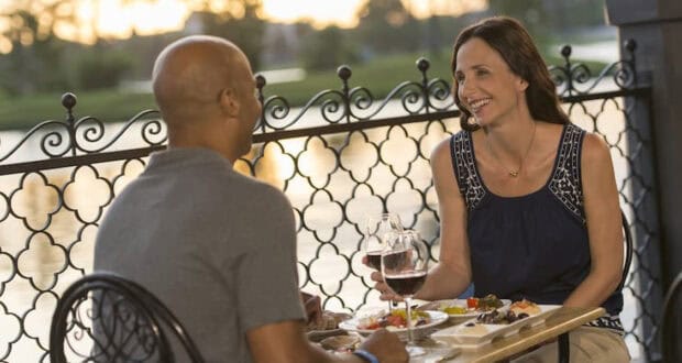 A man and woman sit at an outdoor restaurant table by a decorative railing, sharing Epcot alcoholic drinks, smiling and engaged in conversation as the sun sets over the water in the background.