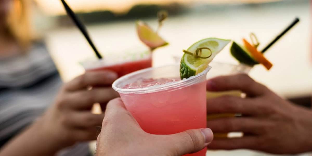 Three people clink colorful Epcot alcoholic drinks in clear plastic cups, each cocktail garnished with lime or orange slices, against a blurred outdoor background.